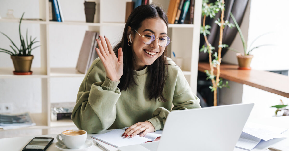 Jovem mulher sorridente com cabelos longos e óculos, usando um moletom verde, participando de uma videochamada em um notebook. Ela está sentada em uma mesa branca, acenando para a tela, com uma xícara de café, celular, caderno aberto e caneta ao seu lado. Ao fundo, há uma estante branca com livros e plantas decorativas, além de uma janela que deixa entrar luz natural (videochamada trabalho híbrido trabalho remoto)