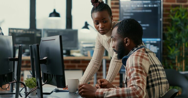 Dois programadores colaborando em um ambiente de escritório moderno. A mulher está em pé ao lado do colega, discutindo um projeto exibido no monitor do computador. O ambiente apresenta uma decoração industrial com detalhes em tijolo aparente e plantas, refletindo um espaço de trabalho colaborativo e tecnológico (trabalho, cultura, time, pessoas, OutSystems, programadores, desenvolvedores, oportunidades, tecnologia, salarios, salario)
