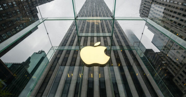 Fachada de uma loja da Apple com o icônico logotipo iluminado em destaque no centro de uma estrutura de vidro. O ângulo da imagem oferece uma perspectiva imponente, mostrando edifícios altos ao fundo e um céu nublado, ressaltando a modernidade e sofisticação da marca (apple, lançamento, inovação)