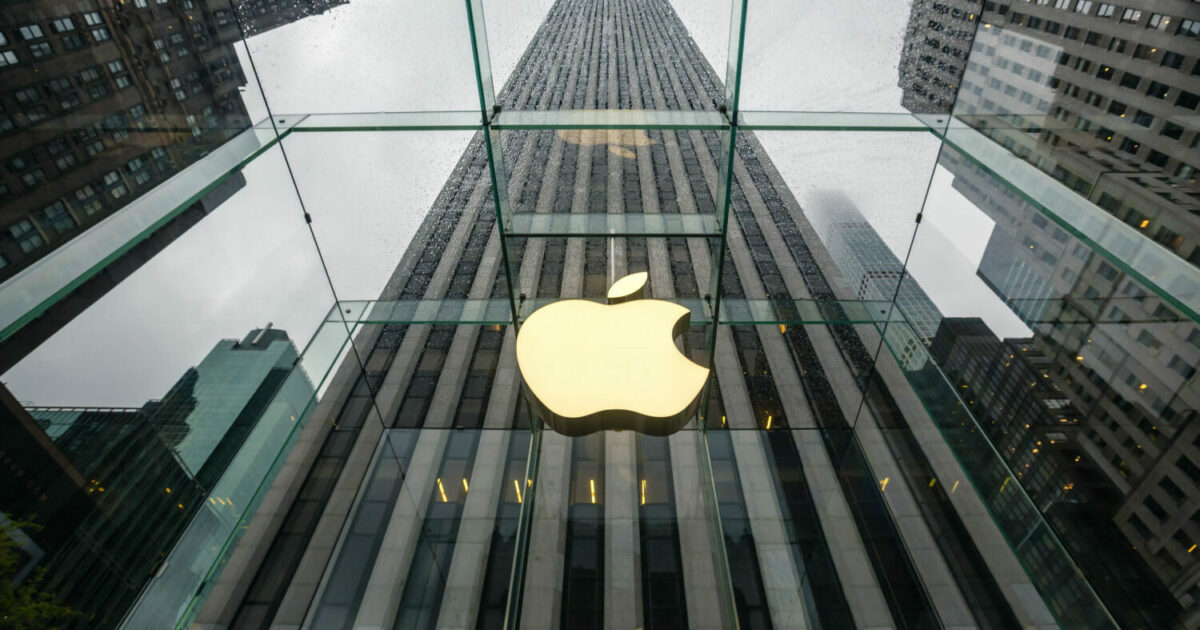 Fachada de uma loja da Apple com o icônico logotipo iluminado em destaque no centro de uma estrutura de vidro. O ângulo da imagem oferece uma perspectiva imponente, mostrando edifícios altos ao fundo e um céu nublado, ressaltando a modernidade e sofisticação da marca (apple, lançamento, inovação)