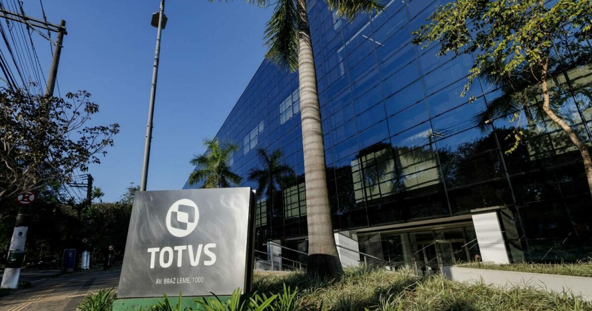 Fachada do prédio da TOTVS, com estrutura moderna de vidro refletindo o céu e as árvores ao redor. Em primeiro plano, destaca-se uma placa com o logotipo da TOTVS e o endereço 'Av. Braz Leme, 1000'. O cenário é complementado por palmeiras e vegetação, transmitindo um ambiente corporativo sofisticado e integrado à natureza (Totvs VTEX Linx)