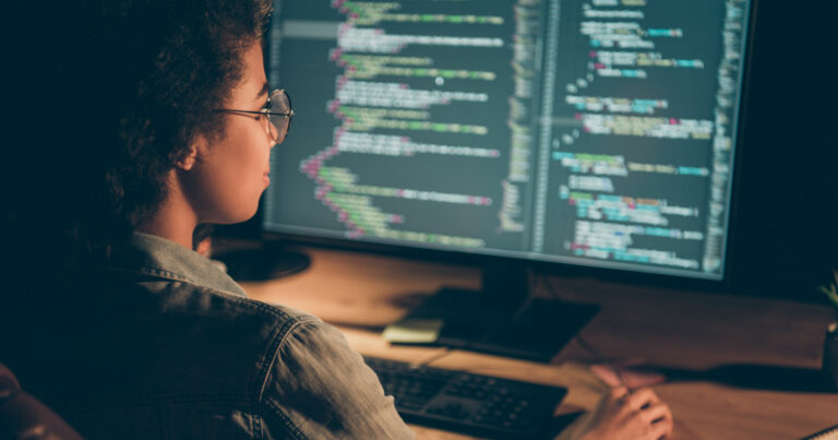 Pessoa trabalhando em programação em um computador, com linhas de código exibidas em uma tela grande. A imagem mostra a profissional de perfil, usando óculos e concentrada no trabalho, em um ambiente de baixa iluminação, o que destaca a tela do monitor. A cena representa o futuro do trabalho em tecnologia e desenvolvimento de software (futuro do trabalho, tecnologia, TI, inteligência artificial, programador, desenvolvedor, emprego, tecnologia, trabalho, empregos, guia salarial, Carreiras na TI, carreira, vida profissiona, Robert Half, salário)