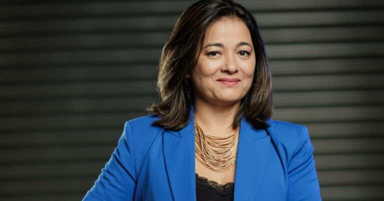 Mulher com cabelos castanhos escuros lisos e pele clara, usando blazer azul, blusa preta rendada e colar dourado com várias voltas. Ela está sorrindo levemente e posando em frente a um fundo escuro com linhas horizontais. Trata-se de Mara Maehara, CIO da Totvs