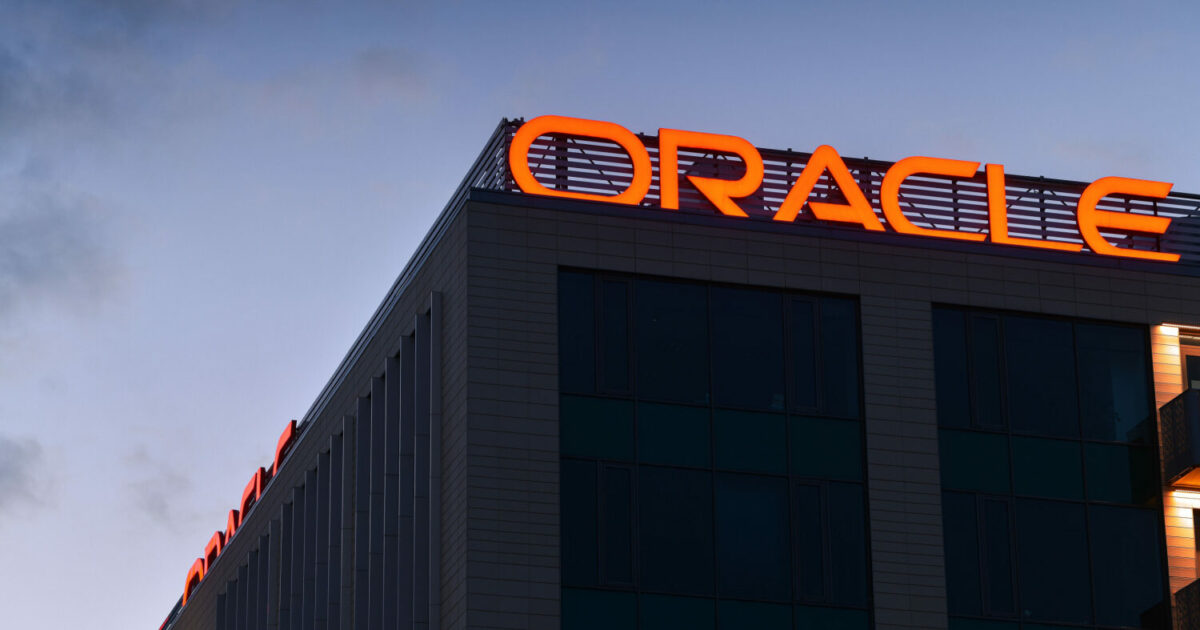 Fachada de um prédio da Oracle ao entardecer, com o logotipo da empresa iluminado em vermelho no topo do edifício. O céu ao fundo está parcialmente nublado, destacando a arquitetura moderna do local (nuvem, Donald Trump)