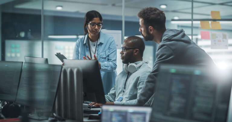 Três profissionais de TI colaborando em frente a vários monitores em um ambiente moderno de segurança cibernética. Uma mulher está em pé com um tablet explicando algo para dois colegas, enquanto um deles está sentado usando fones de ouvido ao redor do pescoço. O ambiente é de alta tecnologia, com luzes suaves e elementos de design contemporâneos (equipes de TI, empresas terceirizadas, cibersegurança)