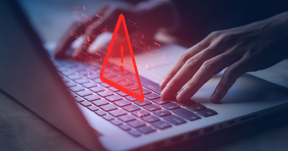 Imagem sombria de um laptop com destaque para um grande ícone vermelho de alerta no centro da tela, simbolizando uma ameaça digital. Há gráficos e elementos visuais em vermelho que remetem a dados e tecnologia, enquanto mãos digitam no teclado, sugerindo um possível ataque cibernético ou atividade maliciosa online (cibersegurança empresas OTRS, empresa)