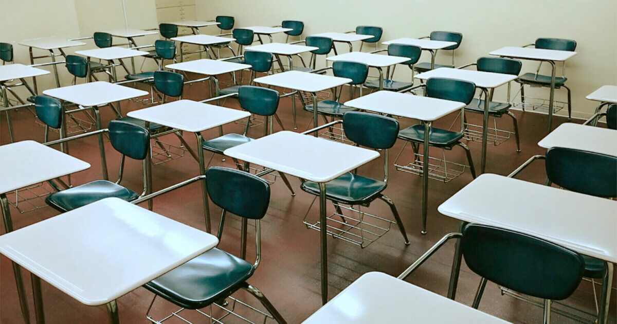 A imagem mostra uma sala de aula vazia, com várias carteiras de madeira organizadas em fileiras, todas acompanhadas de cadeiras também de madeira. Ao fundo, há um grande quadro verde pendurado na parede. O ambiente é bem iluminado por janelas amplas à direita, que deixam a luz natural entrar. As paredes têm um tom claro, contrastando com o revestimento de madeira na parte inferior. A sala transmite uma atmosfera clássica e organizada (Salas de aulas presenciais vazias e online lotadas: EAD vem provocando mudanças na educação)