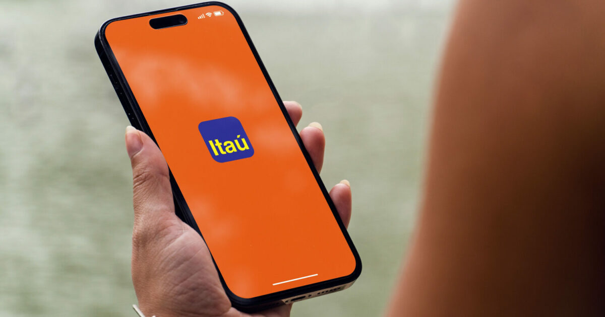Pessoa segurando um smartphone com a tela exibindo o logotipo do Itaú sobre um fundo laranja. A imagem destaca o acesso digital aos serviços do banco em um ambiente externo com fundo desfocado (instituto)