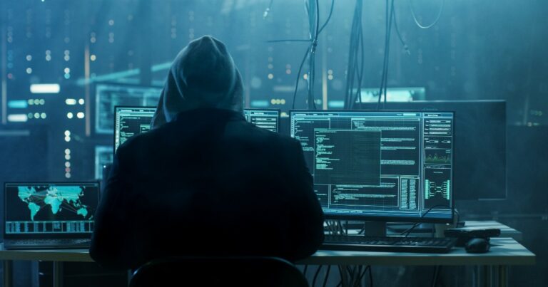 Indivíduo sentado em frente a múltiplas telas de computador em um ambiente escuro, com códigos e mapas globais exibidos nas telas, representando atividades de hacking ou segurança cibernética (cibercriminosos, hacker, cibercrimes, hp crimes cibernéticos, setor financeiro, finanças, bancos, dados, man-in-the-middle)