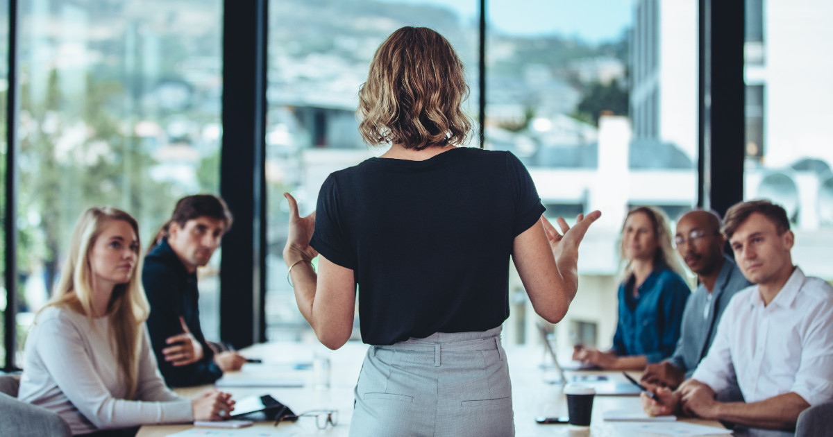 Uma mulher de costas, em pé, liderando uma reunião em um ambiente corporativo moderno. Ela está falando com um grupo de profissionais atentos, sentados ao redor de uma mesa de reunião, com janelas grandes e vista urbana ao fundo, transmitindo um ambiente de liderança e colaboração (liderança, líder, executiva, carreira, reunião, executivo, CEO, CTO, CIO)