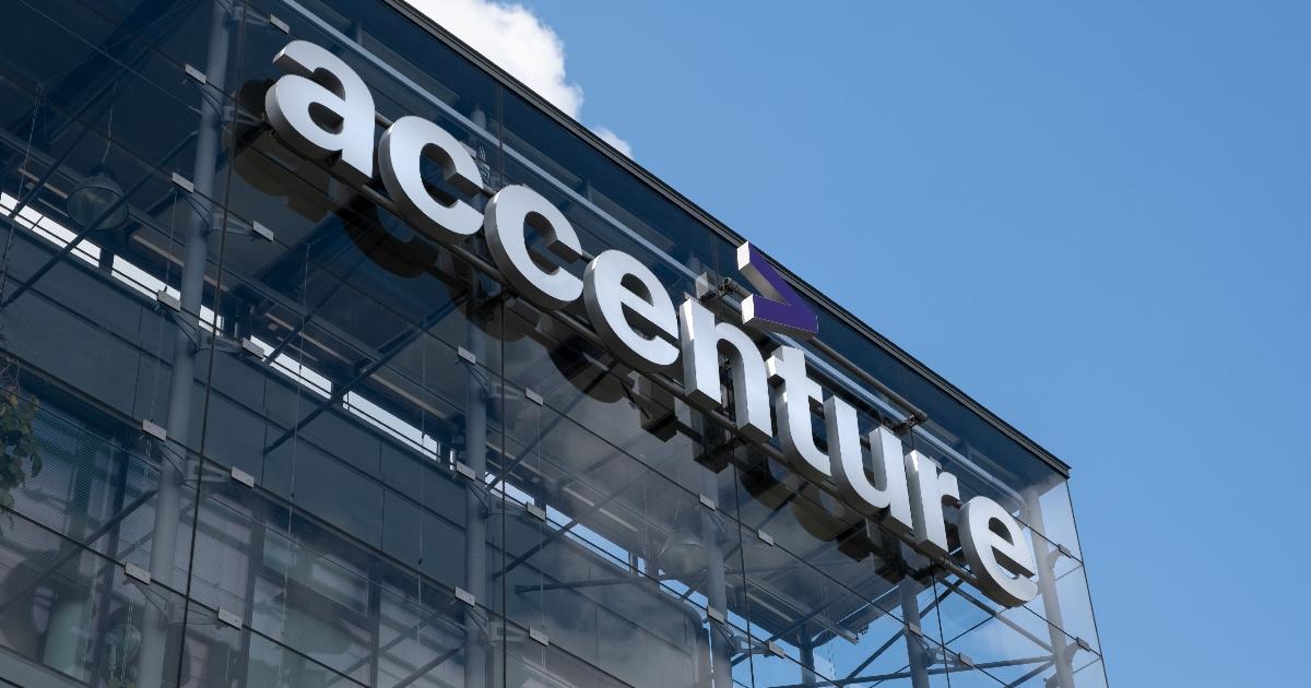 A imagem mostra a fachada de um edifício moderno com a logo da Accenture em destaque. O nome da empresa está escrito em letras brancas, com um acento circunflexo roxo acima da letra "t". A estrutura do prédio é de vidro e metal, refletindo o céu azul com algumas nuvens ao fundo (Accenture, inteligência artificial, aaru, europa)