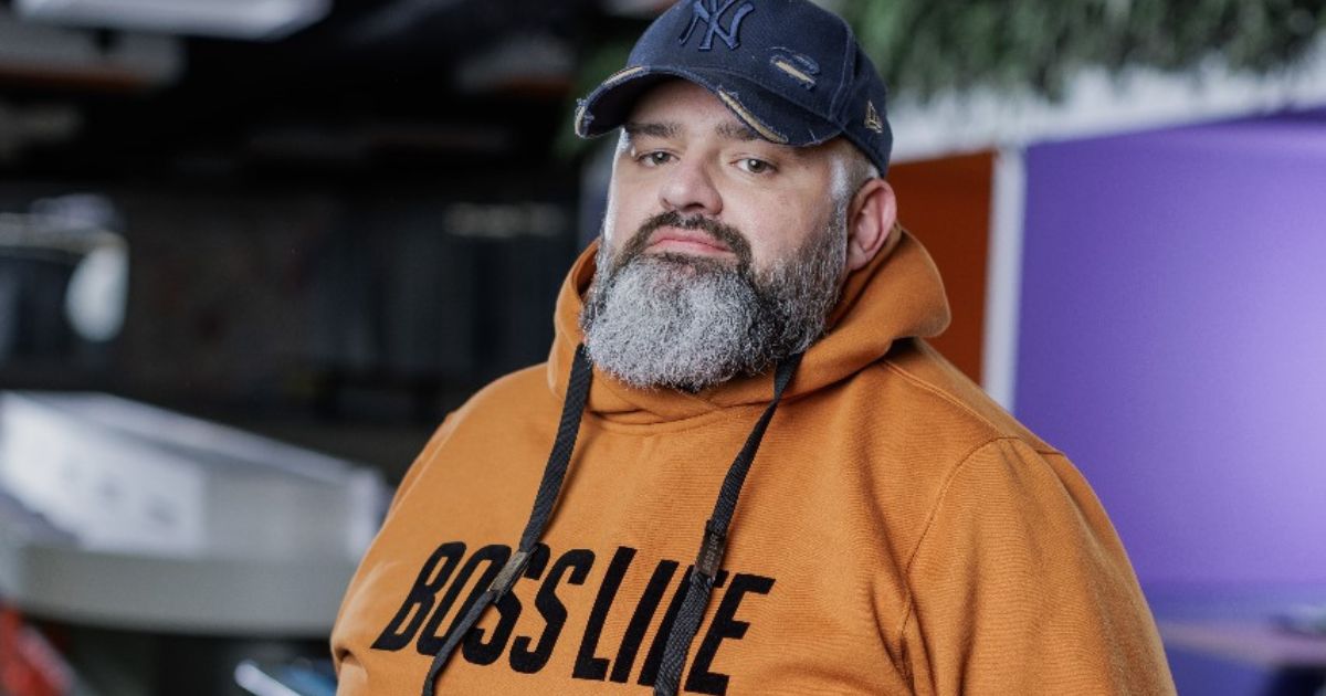 A imagem mostra Joel Backschat, vice-presidente de Tecnologia e Pessoas da FCamara. Ele está com uma expressão séria, usando um boné azul e um moletom laranja com a palavra "BOSSLINE" estampada em letras grandes. A imagem é tirada em um ambiente moderno, com um fundo suave, destacando o roxo. Joel transmite uma postura confiante e profissional Joel Backschat, vice-presidente de Tecnologia e Pessoas da FCamara