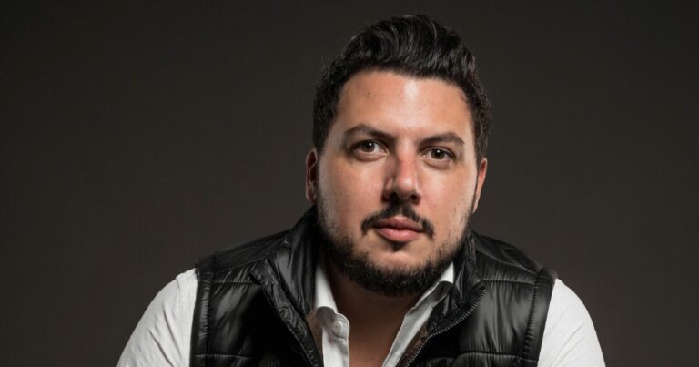 Retrato de Renan Torres, VP comercial da Arklok, com cabelo curto e barba, vestindo uma camisa branca e um colete preto acolchoado. Ele olha diretamente para a câmera com uma expressão séria, em um fundo cinza escuro que destaca seu rosto e vestimenta (arklok, Renan Torres, VP comercial da Arklok, invest)