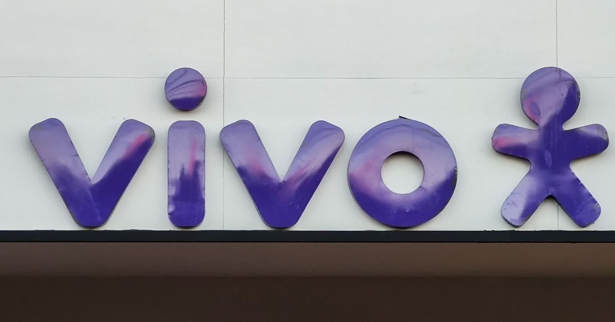 Logotipo da operadora Vivo em destaque, com letras e símbolo na cor roxa fixados em uma parede branca. O design apresenta o nome 'vivo' acompanhado de uma figura estilizada de um boneco, clientes (banco bv)
