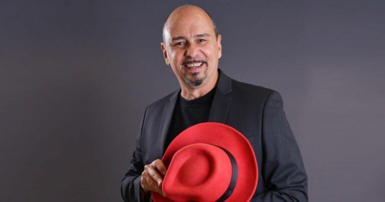 Na imagem, Gilson Magalhães, da Red Hat América Latina, tem uma expressão amigável, vestindo um blazer preto sobre uma camiseta da mesma cor. Ele segura um chapéu vermelho em uma das mãos, com um leve sorriso no rosto. O fundo é neutro, em um tom de cinza escuro, o que destaca ainda mais as cores da vestimenta e do chapéu. O chapéu vermelho é o ponto focal da imagem, adicionando um toque de cor vibrante que contrasta com o traje escuro. A pose e a expressão transmitem um ar confiante e acessível.