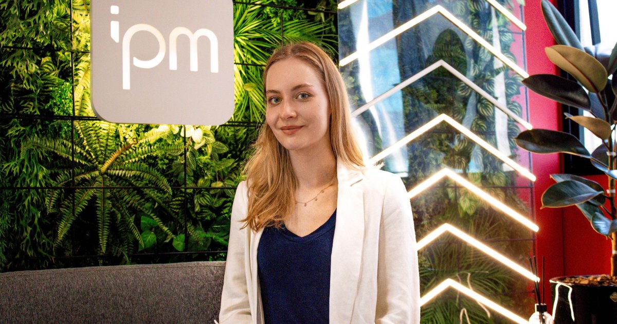 Ana Luiza Mees, head de inteligência artificial da IPM Sistemas, posando em um ambiente moderno e iluminado. Ao fundo, há uma parede verde com plantas e o logotipo da IPM, além de elementos decorativos com luzes geométricas. A imagem transmite inovação e sofisticação, laboratório