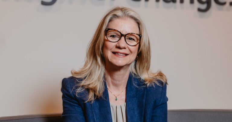 Andrea Klevenhusen, CEO da Ricoh Brasil, sorrindo enquanto está sentada. Ela usa um blazer azul e óculos de armação grande com cabelo loiro comprido e solto. O fundo é desfocado com um texto parcialmente visível