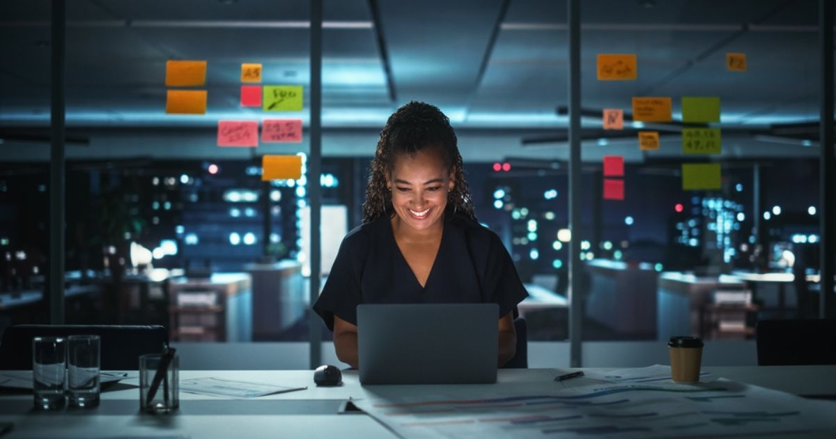 A imagem mostra uma mulher sorridente, trabalhando em um laptop em um ambiente de escritório moderno à noite. Ela está sentada em uma mesa cercada por documentos e copos de vidro, com post-its coloridos colados em um vidro ao fundo, que revela uma vista da cidade iluminada. A cena transmite produtividade, concentração e entusiasmo, sugerindo uma profissional dedicada ou uma empreendedora engajada em suas atividades.