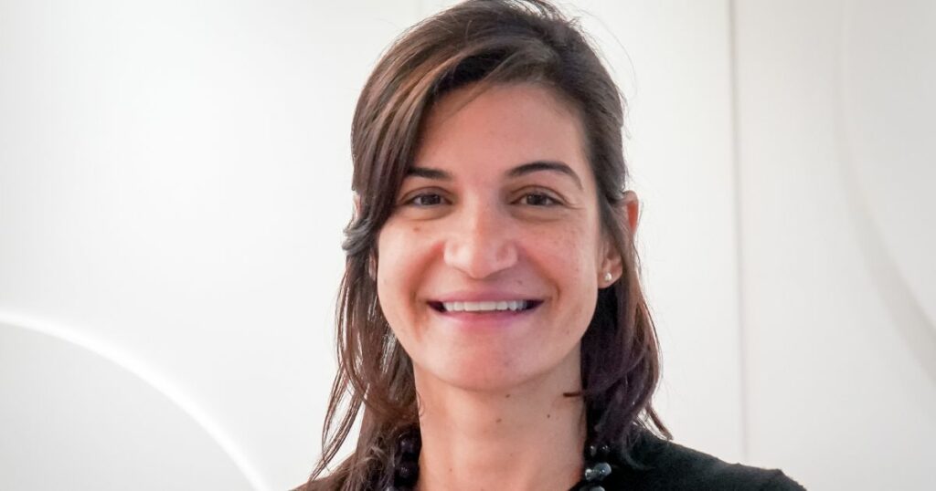 Carolina Maestri, executiva da Odata, sorrindo para a câmera com um fundo neutro e claro. Ela tem cabelos castanhos, usa um colar elegante e transmite uma expressão amigável e acolhedora