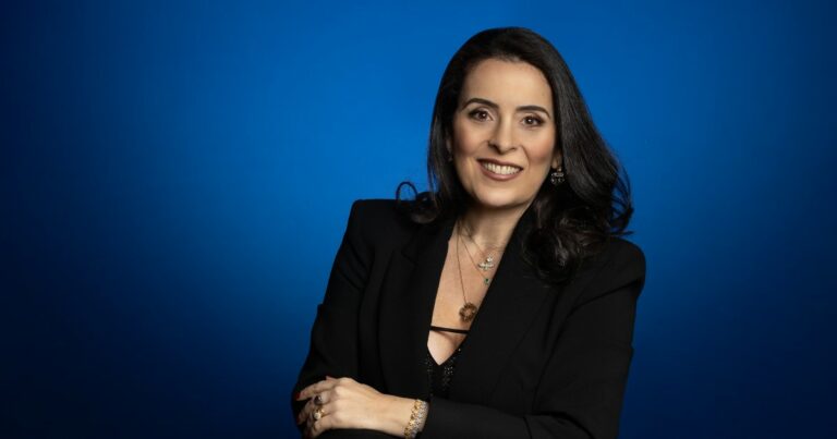Claudia Muchaluat, presidente da Intel Brasil, sorrindo para a câmera com um fundo azul vibrante. Ela está vestida com um blazer preto elegante e usa acessórios delicados, transmitindo uma imagem de confiança e liderança.