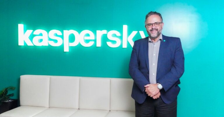 Na imagem, Claudio Martinelli, diretor-geral da Kaspersky para a América Latina, aparece em pé com um semblante amigável, vestindo um terno azul escuro e camisa social clara. Ele está com as mãos entrelaçadas à frente do corpo e usa óculos, posicionado à direita de um sofá branco em um ambiente de escritório. Ao fundo, vê-se uma parede verde com o logotipo da Kaspersky em letras brancas. A imagem transmite profissionalismo e uma atmosfera acolhedora no ambiente da empresa.