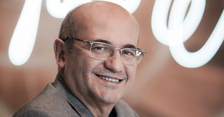 Cleber Morais, diretor-geral de vendas da AWS no Brasil, sorrindo para a câmera com um fundo desfocado em tons claros. Ele está usando óculos elegantes e um paletó cinza, transmitindo uma imagem de confiança e simpatia (IA generativa)