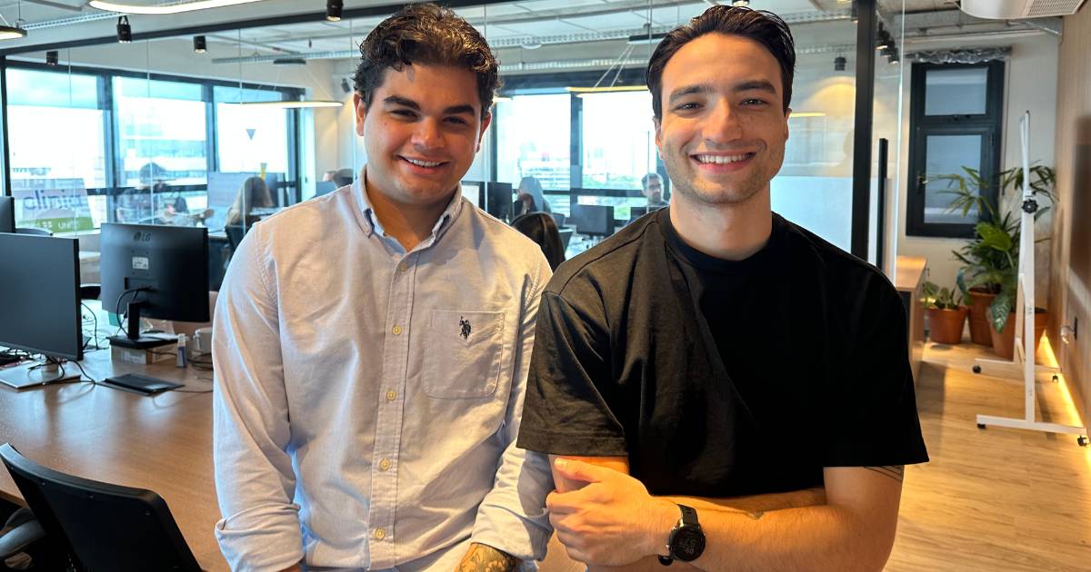 Leonardo Bork e Artur Rossi, fundadores da Clinia, posando em um ambiente moderno de escritório. Leonardo veste uma camisa social azul clara, enquanto Artur usa uma camiseta preta casual. Ao fundo, há mesas de trabalho com computadores, janelas amplas e decoração contemporânea, refletindo o espírito inovador da startup