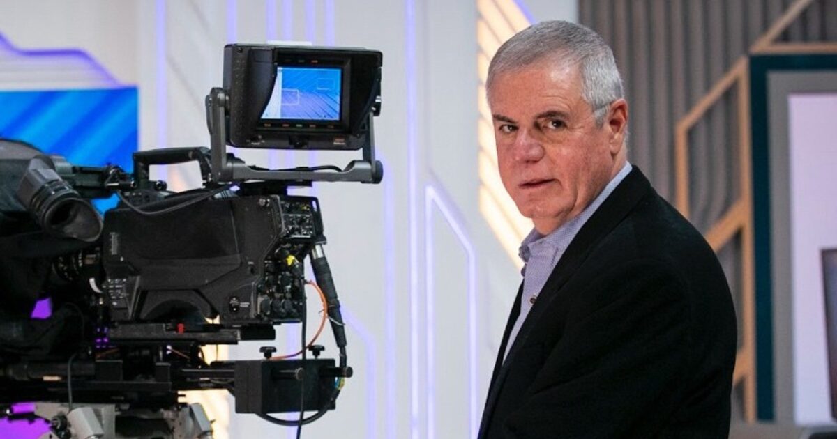 Alfonso Aurin, novo superintendente de Distribuição, Tecnologia & Serviços do SBT, em um estúdio de televisão ao lado de uma câmera profissional, olhando para a câmera com uma expressão séria. O ambiente tem iluminação moderna e elementos tecnológicos ao fundo