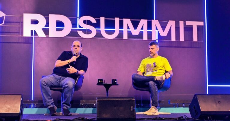 Foto de Germán Quiroga, conselheiro e investidor, sócio da Omini11 Consultoria, à esquerda, segurando um microfone enquanto fala durante o RD Summit. Ao seu lado direito, Patrick Scripilliti, especialista de digital commerce da Totvs, veste uma camiseta amarela e escuta atentamente. O letreiro iluminado 'RD Summit' e luzes de neon criam uma atmosfera vibrante no palco do evento, e-commerce