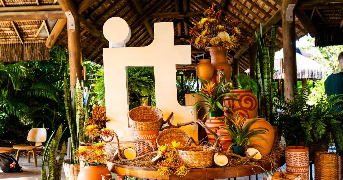A imagem mostra uma decoração colorida e vibrante em um evento, com destaque para as letras "IT", de IT Forum, em grande tamanho, que parecem ser um elemento central. A letra está rodeada por vasos de cerâmica com desenhos decorativos, cestas de vime e plantas tropicais, como bromélias e samambaias. O ambiente é rústico, com uma estrutura de palha ao fundo, sugerindo um espaço aberto ou ao ar livre. A composição transmite uma sensação de acolhimento e natureza, ideal para eventos ou encontros temáticos.