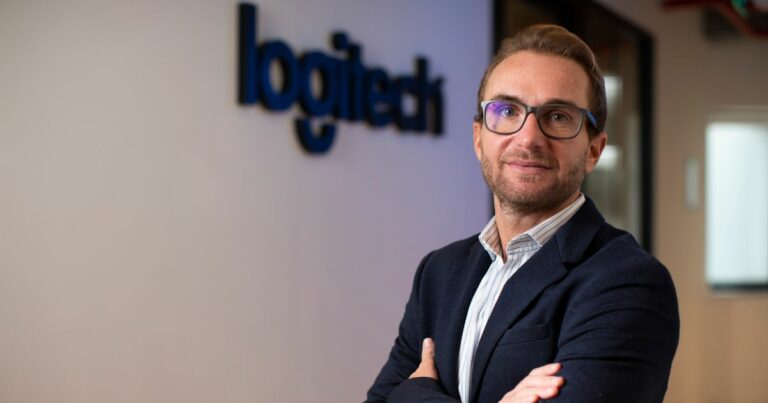 A imagem mostra um homem de óculos e barba rala, vestindo um blazer azul escuro e camisa listrada, posicionado de braços cruzados. Ao fundo, parcialmente desfocado, está o logotipo da Logitech na parede. Ele tem uma expressão séria, confiante e está em um ambiente corporativo, possivelmente o escritório da Logitech. O executivo é o Jairo Rozenblit, CEO da Logitech.