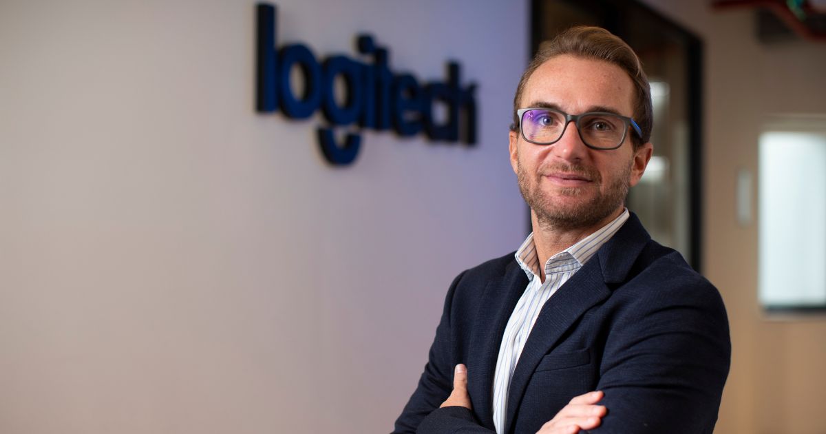 A imagem mostra um homem de óculos e barba rala, vestindo um blazer azul escuro e camisa listrada, posicionado de braços cruzados. Ao fundo, parcialmente desfocado, está o logotipo da Logitech na parede. Ele tem uma expressão séria, confiante e está em um ambiente corporativo, possivelmente o escritório da Logitech. O executivo é o Jairo Rozenblit, CEO da Logitech.