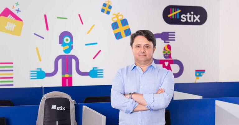 Jorge Proença, CIO da Stix, posando em um ambiente de escritório moderno, com uma parede decorada com ilustrações coloridas que representam diferentes serviços e atividades. Ele está de braços cruzados, vestindo uma camisa de botão azul clara, com uma expressão séria e confiante. Ao lado dele, há uma mochila cinza com o logotipo da Stix