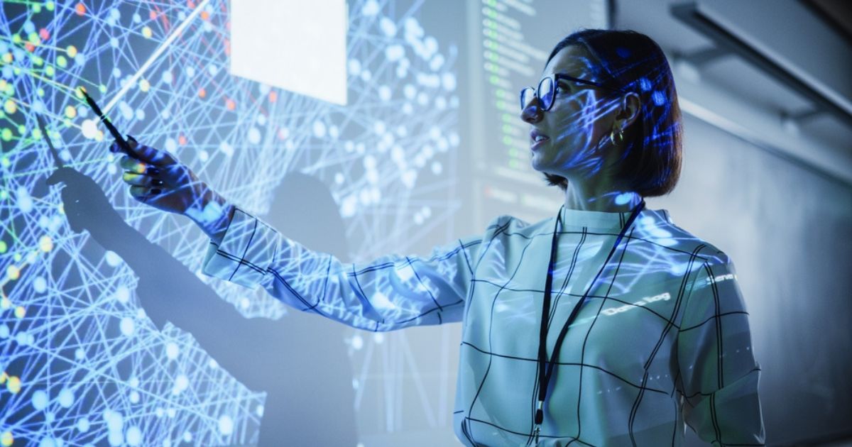 A imagem mostra uma mulher em um ambiente educacional ou profissional, apresentando informações relacionadas à tecnologia. Ela está apontando para uma projeção digital que exibe uma rede interconectada de nós, sugerindo temas como inteligência artificial, big data ou análise de redes. A mulher usa óculos, veste uma camisa com padrão geométrico, e parece engajada enquanto explica ou destaca algo na projeção, reforçando uma atmosfera de inovação e aprendizado tecnológico. A iluminação cria uma interação visual interessante entre o conteúdo projetado e a pessoa, simbolizando a fusão entre o humano e a tecnologia.
