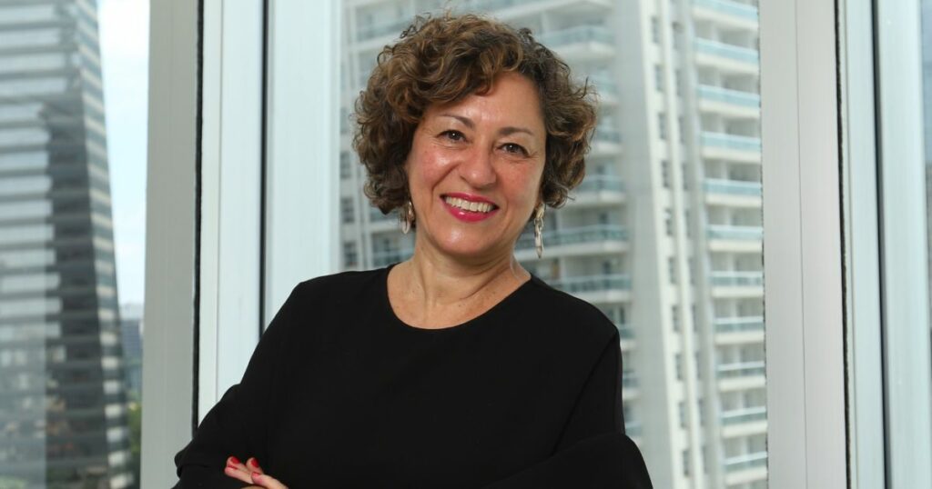 Nelmara Arbex, executiva da KPMG, posando sorridente em um ambiente corporativo. Ela veste uma blusa preta elegante, com o fundo mostrando janelas de vidro que revelam prédios modernos e uma vista urbana ao longe