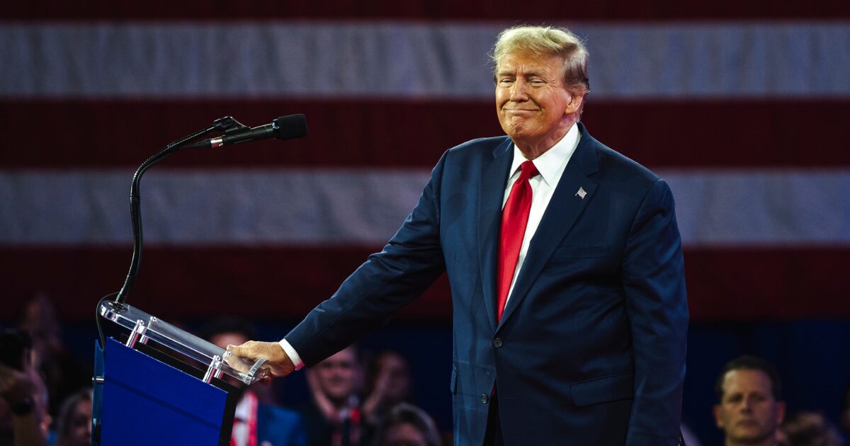 Imagem de Donald Trump de terno azul-escuro e gravata vermelha em pé, ao lado de um púlpito com microfone. Ele está com uma expressão de leve sorriso e um fundo de bandeira dos Estados Unidos em destaque.