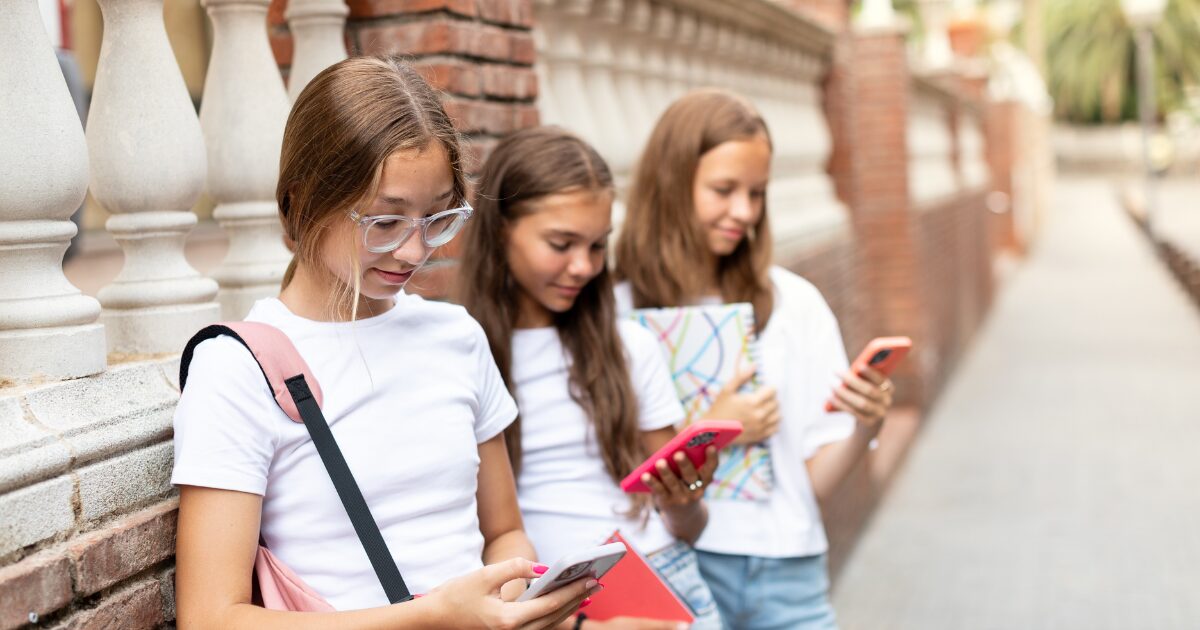 Três adolescentes em pé ao lado de uma parede de tijolos e balaústres estão olhando para seus celulares. Elas vestem camisetas brancas e seguram celulares coloridos, aparentemente imersas em redes sociais ou mensagens. Uma das meninas carrega uma mochila rosa no ombro e óculos de armação transparente, enquanto outra segura um caderno com capa colorida. A cena reflete o uso frequente das redes sociais entre jovens e sua conexão com a tecnologia em espaços urbanos.