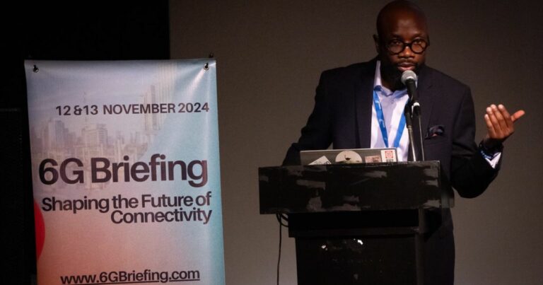 A imagem mostra um homem em pé, vestido com um fato azul com padrão axadrezado, camisa branca e gravata azul-escura. Ele usa óculos redondos e possui barba. Ao fundo, há um cartaz azul com texto em português que destaca a visão e valores de uma instituição, mencionando palavras como ética, transparência, inovação e responsabilidade socioambiental. O homem está posicionado ao lado do cartaz, transmitindo um ar profissional e confiante. 6G