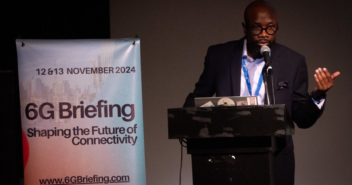 A imagem mostra um homem em pé, vestido com um fato azul com padrão axadrezado, camisa branca e gravata azul-escura. Ele usa óculos redondos e possui barba. Ao fundo, há um cartaz azul com texto em português que destaca a visão e valores de uma instituição, mencionando palavras como ética, transparência, inovação e responsabilidade socioambiental. O homem está posicionado ao lado do cartaz, transmitindo um ar profissional e confiante. 6G