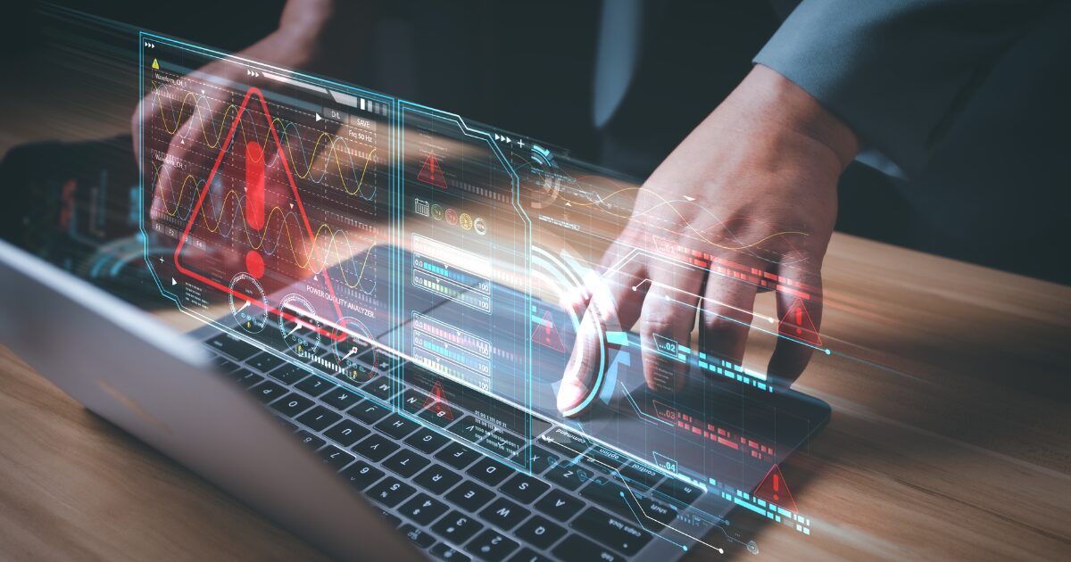 A imagem apresenta um laptop com a tela exibindo gráficos holográficos avançados e elementos digitais flutuantes. Um grande ícone de alerta vermelho com um ponto de exclamação ocupa o centro, indicando um possível erro ou situação crítica. Ao fundo, há gráficos de ondas, indicadores de desempenho e barras de progresso coloridas. As mãos de uma pessoa, vestindo roupa formal, interagem com o teclado, sugerindo um ambiente de análise tecnológica ou de gestão de sistemas críticos. A composição transmite uma sensação de alta tecnologia e monitorização em tempo real. (engenharia social, Netwrix, ICTS)