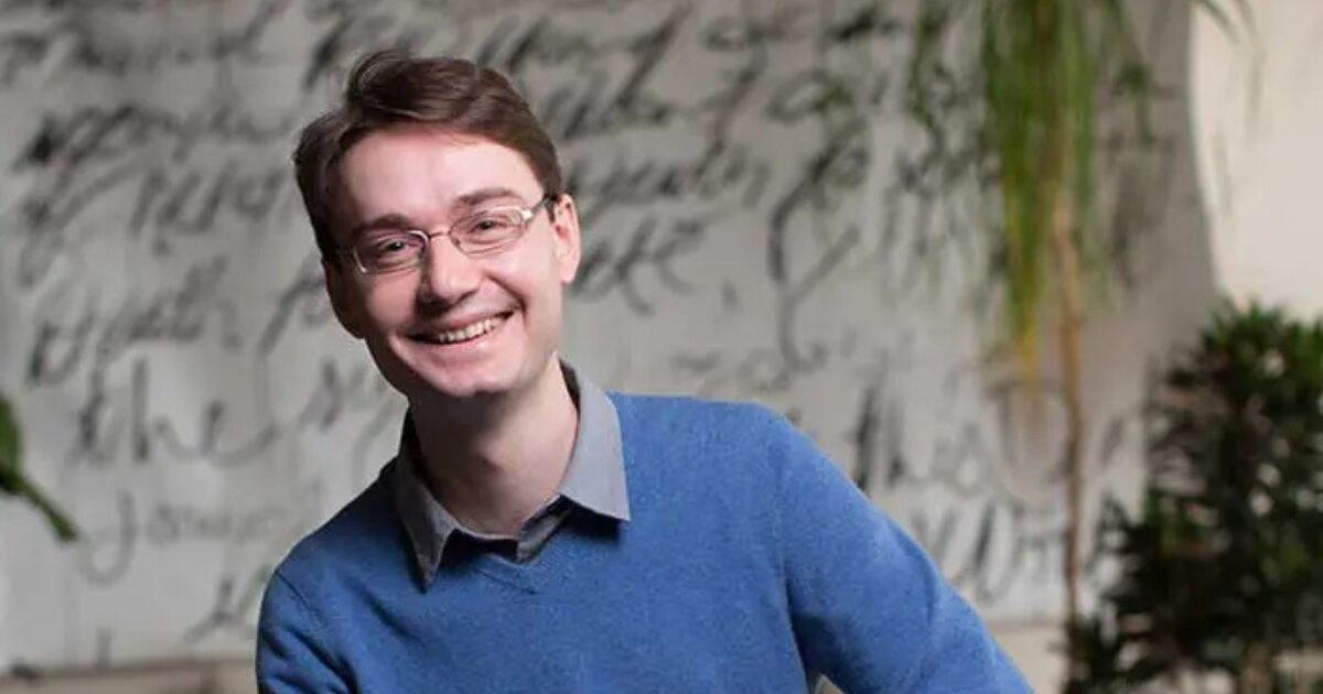 François Chollet usando óculos de aro fino e uma camisa azul, está sorrindo para a câmera. O fundo é artístico, com uma parede coberta por textos manuscritos e plantas, dando um tom informal e criativo à imagem. Ele parece relaxado e amigável, com um visual que sugere um ambiente profissional descontraído.