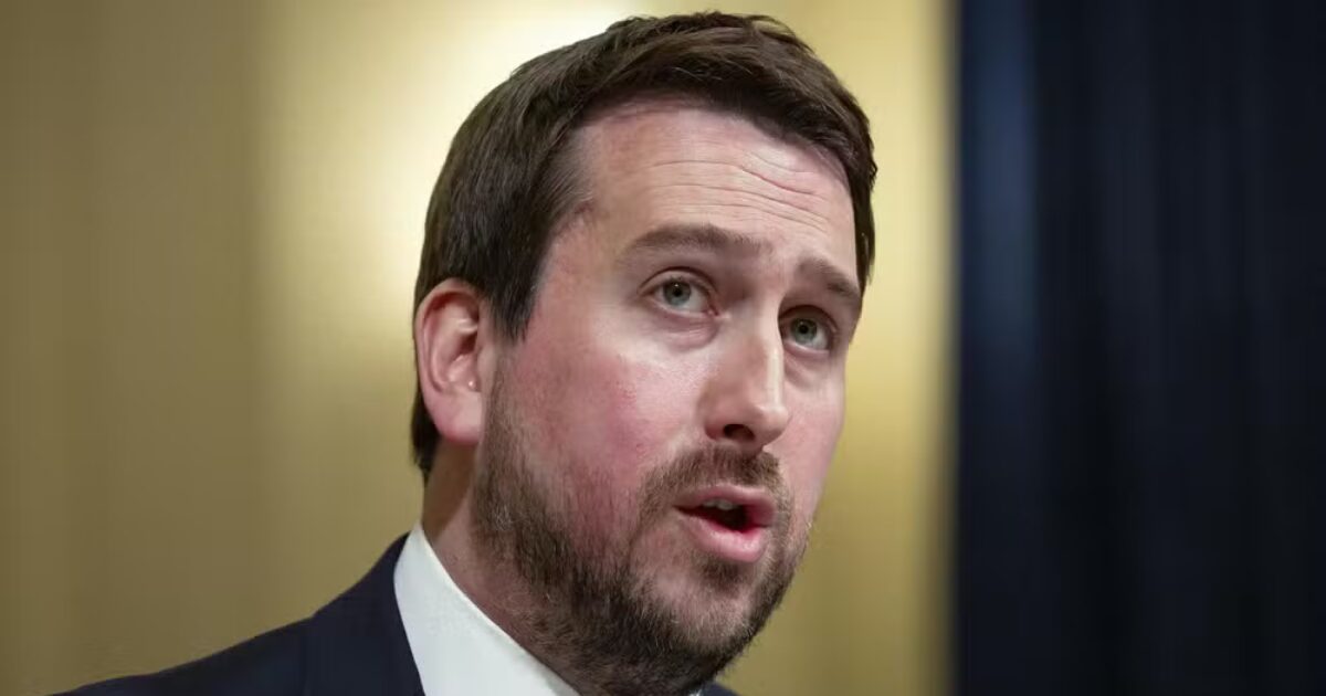 A imagem mostra Nick Pickles com cabelos curtos e barba curta, usando um terno escuro e gravata, falando enquanto olha ligeiramente para o lado. O fundo é neutro, com iluminação suave, destacando o rosto da pessoa na imagem.