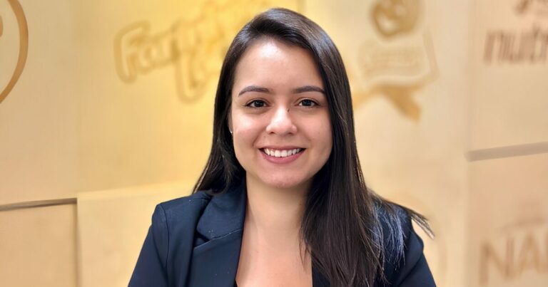 A imagem mostra uma mulher sorridente, vestida de forma profissional, com um blazer preto. Ela tem cabelo longo e escuro, pele clara e está em um ambiente que parece ser corporativo, com o logo da Nestlé ao fundo, sugerindo que ela é uma gerente da empresa.