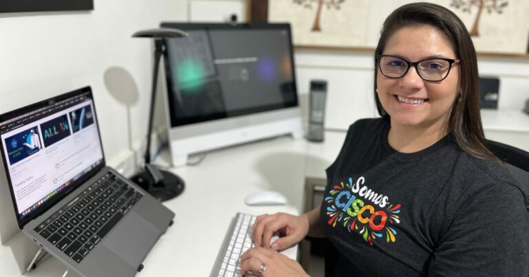 Mulher sorridente trabalhando em um ambiente home office, usando uma camiseta preta com a frase "Somos Cisco" estampada. À sua frente, um teclado e dois monitores: um laptop aberto com informações da empresa e um computador de mesa ao fundo. O ambiente é iluminado e organizado, com detalhes decorativos na parede ao fundo.