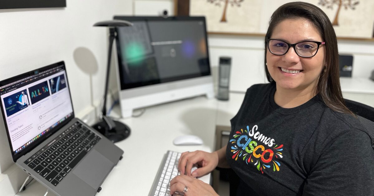 Mulher sorridente trabalhando em um ambiente home office, usando uma camiseta preta com a frase "Somos Cisco" estampada. À sua frente, um teclado e dois monitores: um laptop aberto com informações da empresa e um computador de mesa ao fundo. O ambiente é iluminado e organizado, com detalhes decorativos na parede ao fundo.