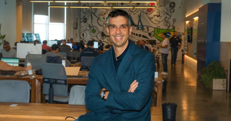 Foto de Ricardo Brandão, CEO da SkyOne, sorrindo enquanto está em pé com os braços cruzados em um ambiente de escritório moderno e colaborativo. O espaço ao fundo é movimentado, com várias pessoas trabalhando em mesas compartilhadas, e uma parede decorada com grafites e ilustrações vibrantes, criando uma atmosfera descontraída e criativa
