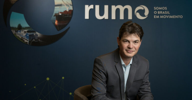 Marco Andriola, diretor de tecnologia da Rumo Logística, sentado em um ambiente corporativo com o logotipo da empresa e o slogan 'Somos o Brasil em Movimento' ao fundo. Ele veste um terno cinza e sorri discretamente, com uma postura profissional