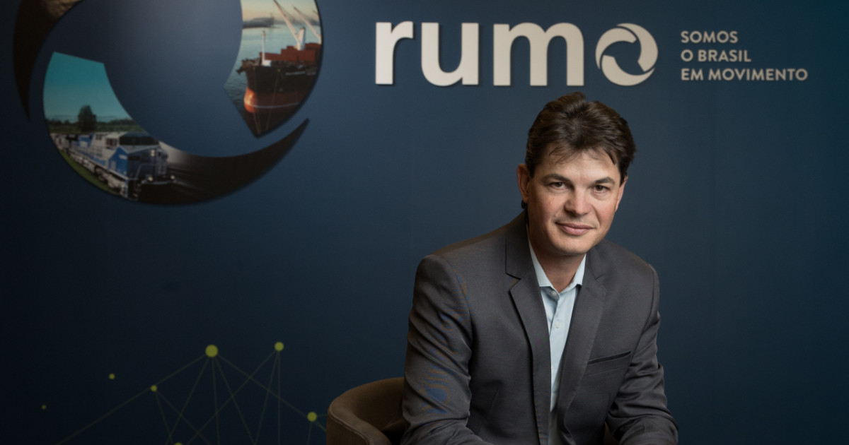 Marco Andriola, diretor de tecnologia da Rumo Logística, sentado em um ambiente corporativo com o logotipo da empresa e o slogan 'Somos o Brasil em Movimento' ao fundo. Ele veste um terno cinza e sorri discretamente, com uma postura profissional