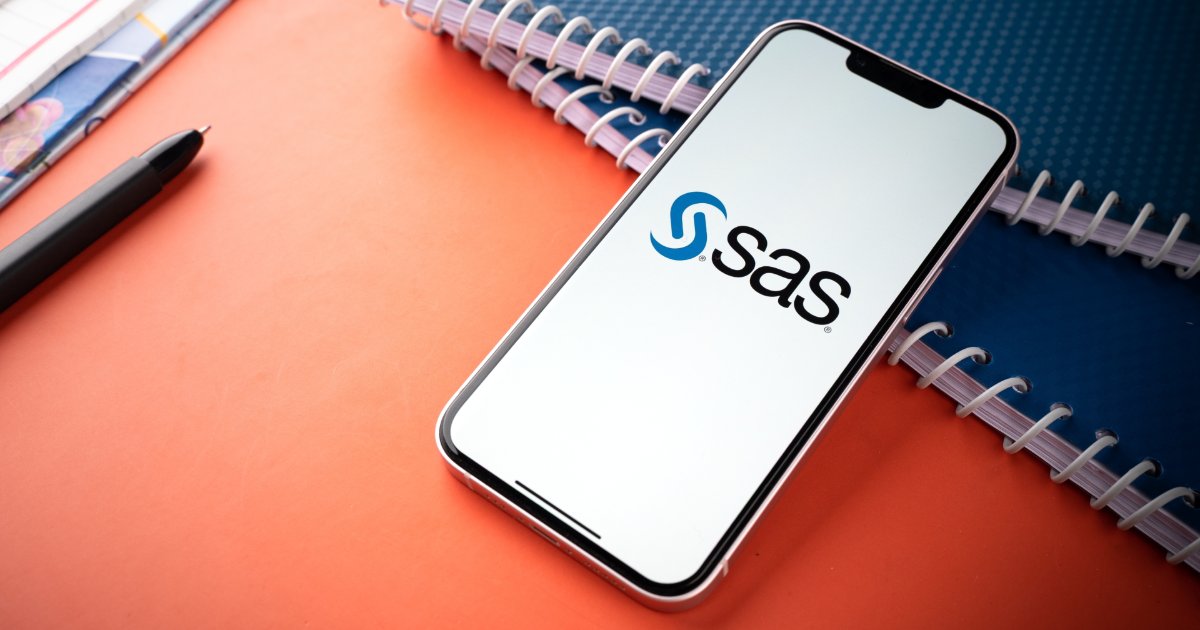Smartphone com o logotipo da SAS exibido na tela, posicionado sobre uma mesa laranja, ao lado de cadernos em espiral e uma caneta preta