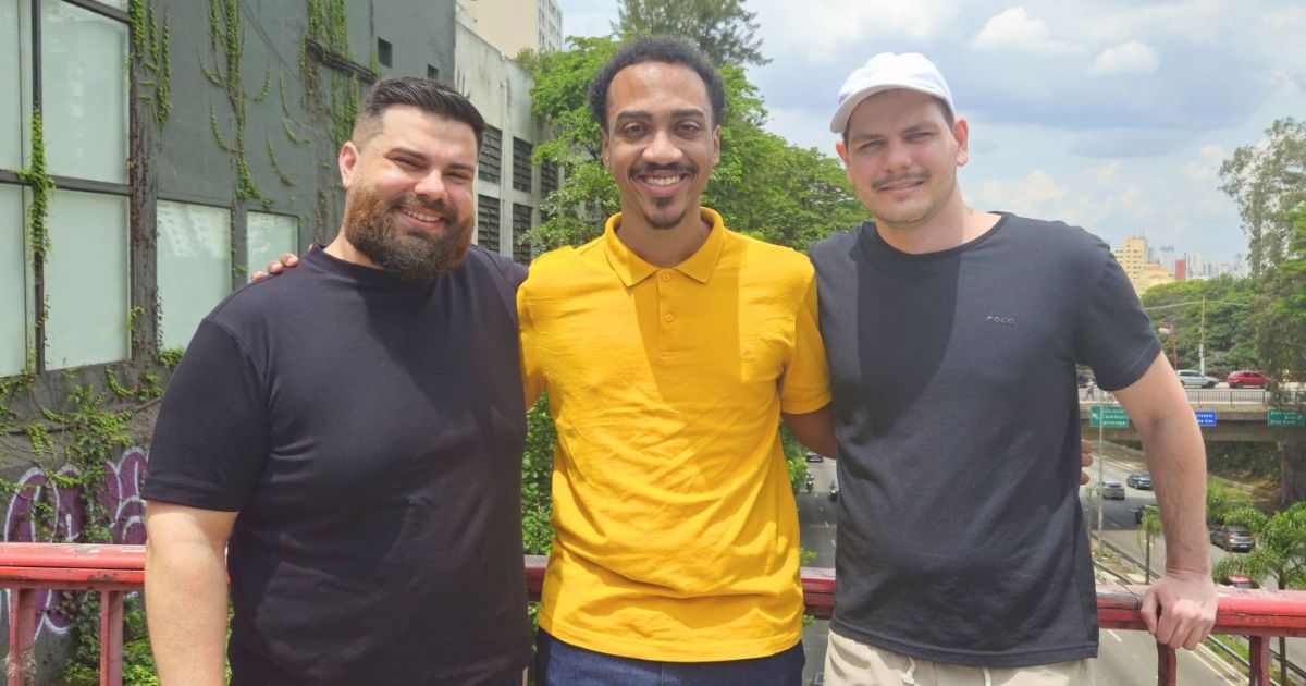 Ivan, Victor e Raphael, da Styleflow, posando juntos ao ar livre. Ivan veste uma camisa preta, Victor usa uma camisa polo amarela e Raphael está com uma camisa preta e boné branco. Eles sorriem para a câmera em frente a um cenário urbano com vegetação e prédios ao fundo